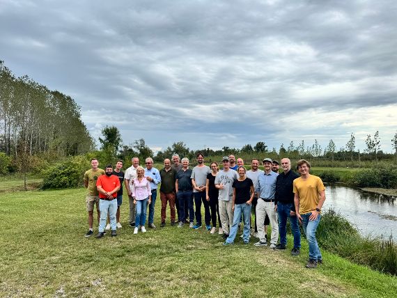 Gruppenfoto neben einem Fluss unter bewölktem Himmel, im Hintergrund Bäume und Sträucher.