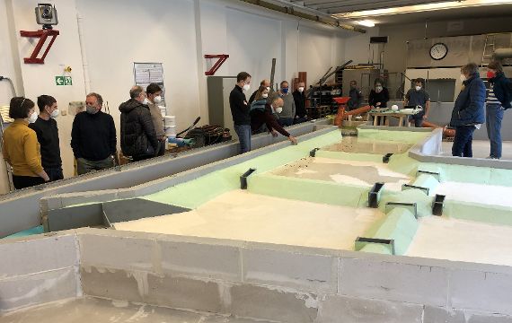 Blick auf das physikalische Modell, das von Besucher:innen umringt ist. Das Modell stellt ein Schwebstoffabsetzbecken dar, welches aus fünf Teilbecken besteht. In den ersten beiden Becken fließt Wasser.