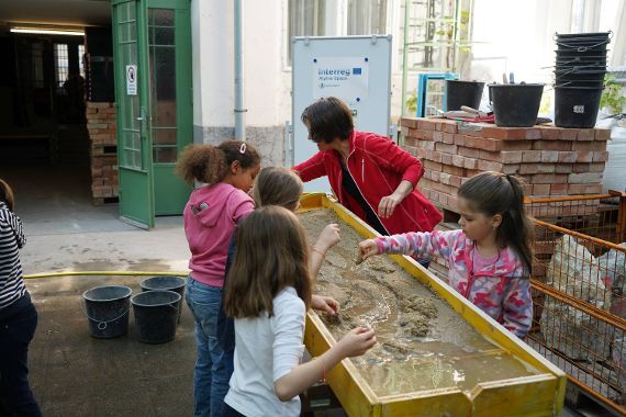 Das Bild zeigt vier Kinder und eine Frau die an einer etwa einen Meter breiten Rinne aus Holz stehen. Die Rinne ist mit Sand gefüllt. Im Sand ist eine Vertiefung, durch die Wasser fließt. Die Personen an der Rinne schauen, wie das fließende Wasser den Sand umlagert und versuchen mit ihren Händen den Wasserstrom zu lenken. Im Hintergrund sieht man eine große grüne Türe. Auf der Seite stehen viele Mauerziegel und Eimer.