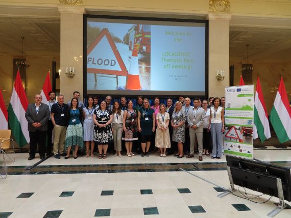 Gruppenbild in der Marmorhalle des ungarischen Innenministeriums. 26 Personen stehen vor der großen Präsentationswand, auf der steht „Welcome to the Localience Thematic Kick off Meeting!“. Links und rechts der Personen sind ungarische Flaggen zu sehen.