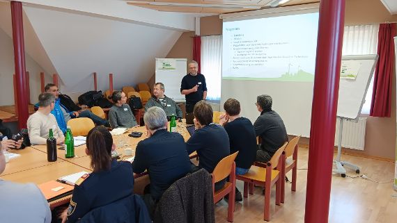 Die Teilnehmenden der Veranstaltung sitzen in einem Sitzungszimmer. Eine Person hält einen Vortrag anhand einer Präsentation.