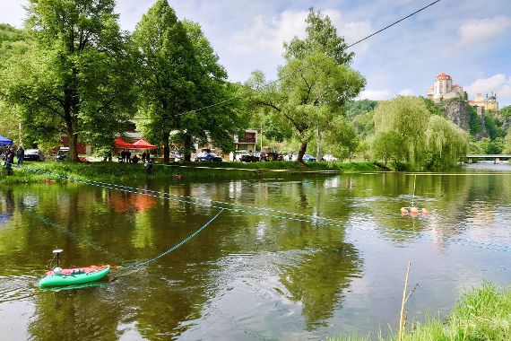 Blick vom Ufer auf einen Fluss, über den zwei Leinen gespannt sind, mit denen Messboote gezogen werden. Im Hintergrund auf einem Felsen das Schloss Vranov nad Dyjí.